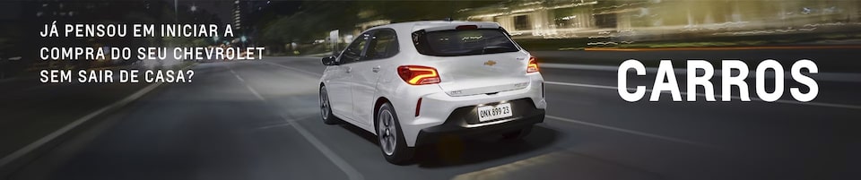 Um carro Chevrolet branco está circulando em uma rua da cidade à noite. O texto em português pergunta se o espectador já pensou em comprar um Chevrolet sem sair de casa, com a palavra CARROS em destaque.