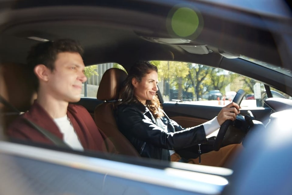 Imagem de um casal dentro de um Chevrolet. A mulher, branca, loira, trajando uma jaqueta de couro preta, dirigindo e com as mãos sobre o volante. No passageiro, um homem bramco, trajando camiseta branca e blazer cor de vinho.