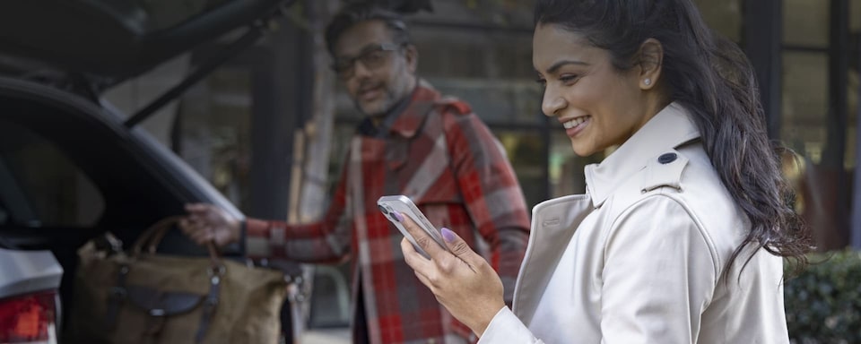 Ao fundo da imagem, um homem branco, de camisa xadrez e óculos, retirando uma mala do porta malas de um Chevrolet. Em primeiro plano, uma mulher branca, jovem, de cabelos presos e jaqueta clara, sorri e olha o celular que segura nas mãos.