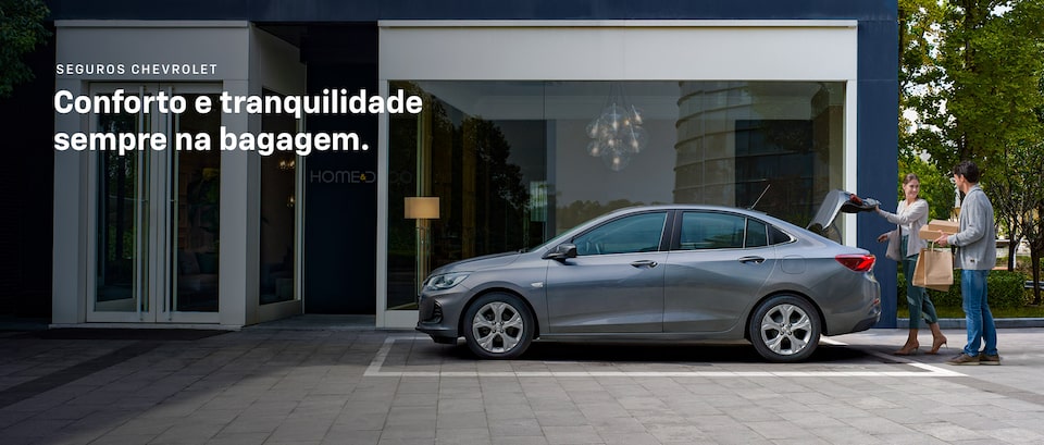 Imagem de um Chevrolet Onix Plus, na cor cinza, estacionado em vaga na rua. Um casal, com compras nas mãos, abrindo o porta malas do carro para guardar as sacolas.