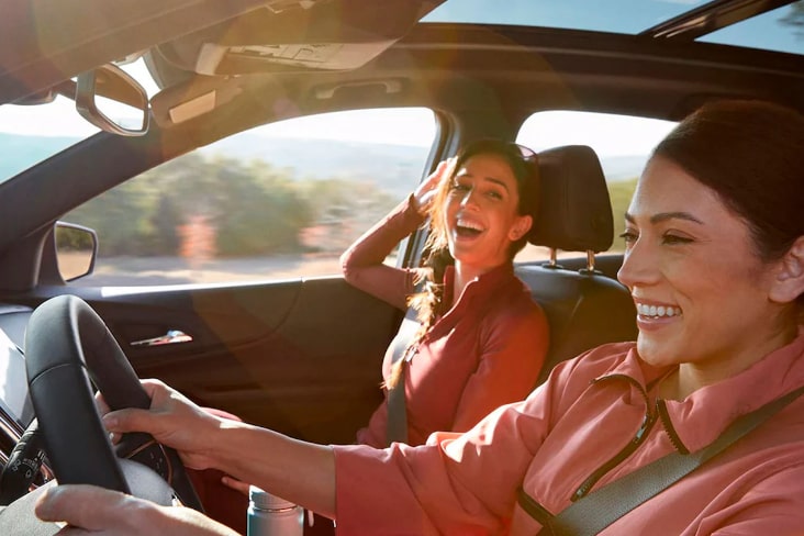 Imagem registrada dentro de um veículo, onde mostra duas mulheres sorrindo e felizes, uma condutora e uma passageira.