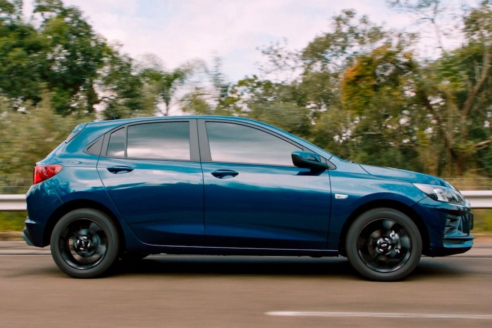 Chevrolet Onix edição comemorativa 100 Anos visto lateralmente em estrada arborizada, com pintura Azul Boreal e rodas escuras.