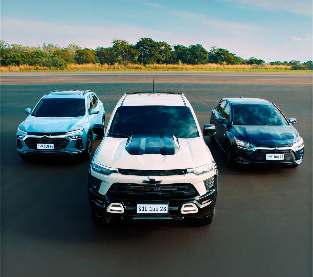 Três veículos Chevrolet alinhados em pista aberta. S10 ao centro, Tracker à esquerda e um Onix à direita, todos em vista frontal.