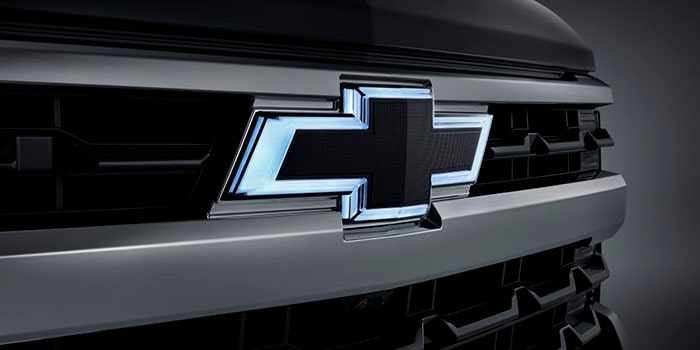 Imagem em close da grade dianteira da nova picape Chevrolet Silverado, preto, dando destaque ao logo Chevrolet iluminado com neon em sua volta.